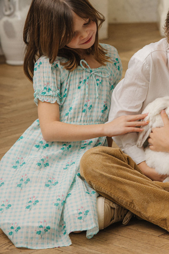 A young girl in a blue floral spring dress sits on the floor, smiling as she gently pets a white rabbit held by another child next to her, who is wearing the Ivy City Co Mini Jessie Dress in Bloom Gingham.