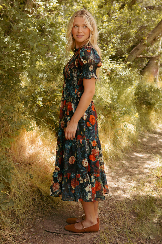 A woman with blonde hair stands on a sunlit forest path, looking over her shoulder as she wears the Cordelia Dress in Moody Floral by Ivy City Co. Surrounded by lush bushes and trees, she completes this maternity-friendly look with brown flats, creating a serene scene.