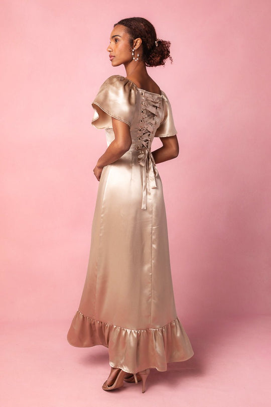 Wearing the Maddie Dress in Champagne Satin by Ivy City Co, a woman stands in profile against a pink background. The dress features a ruffled hem, short sleeves, and a lace-up corset-style back. She completes her elegant and modest bridesmaid look with pearl earrings and heels.