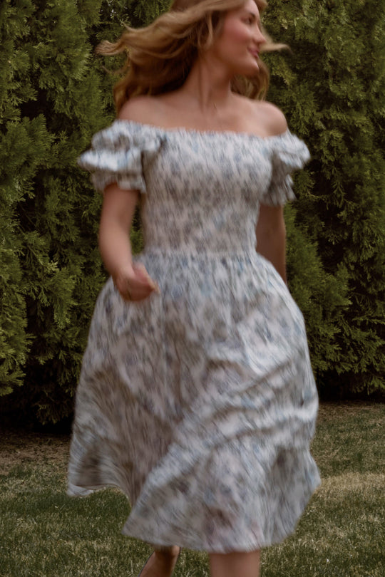 A woman runs outdoors on grass, her hair flowing behind her, wearing the Ivy City Co Valencia Dress in Ivory Climbing Vine Bloom. The off-shoulder, floral design and airy elegance of this maternity-friendly dress are highlighted against a backdrop of green bushes.