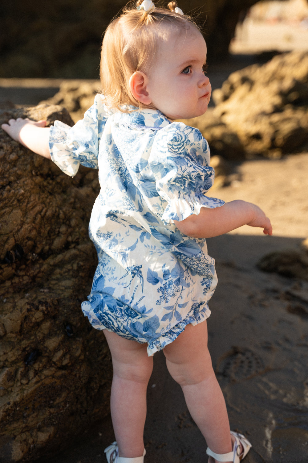 Wearing the Ivy City Co Baby Sophia Romper in Blue Floral, a toddler stands on a sandy beach with one hand resting on a rock, glancing back over their shoulder as sunlight catches their fair hair against the rocky backdrop.