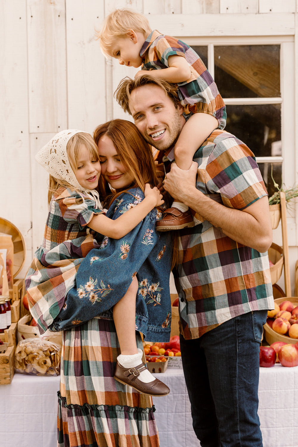 A smiling family of four in matching plaid and floral outfits poses in front of a white wooden wall, with a table of apples and produce behind them. The mother, wearing the Ivy City Co Mini Alice Dress in Maize Gingham featuring a smocked bodice and puff sleeves, hugs their child while the father carries their other child on his shoulders.