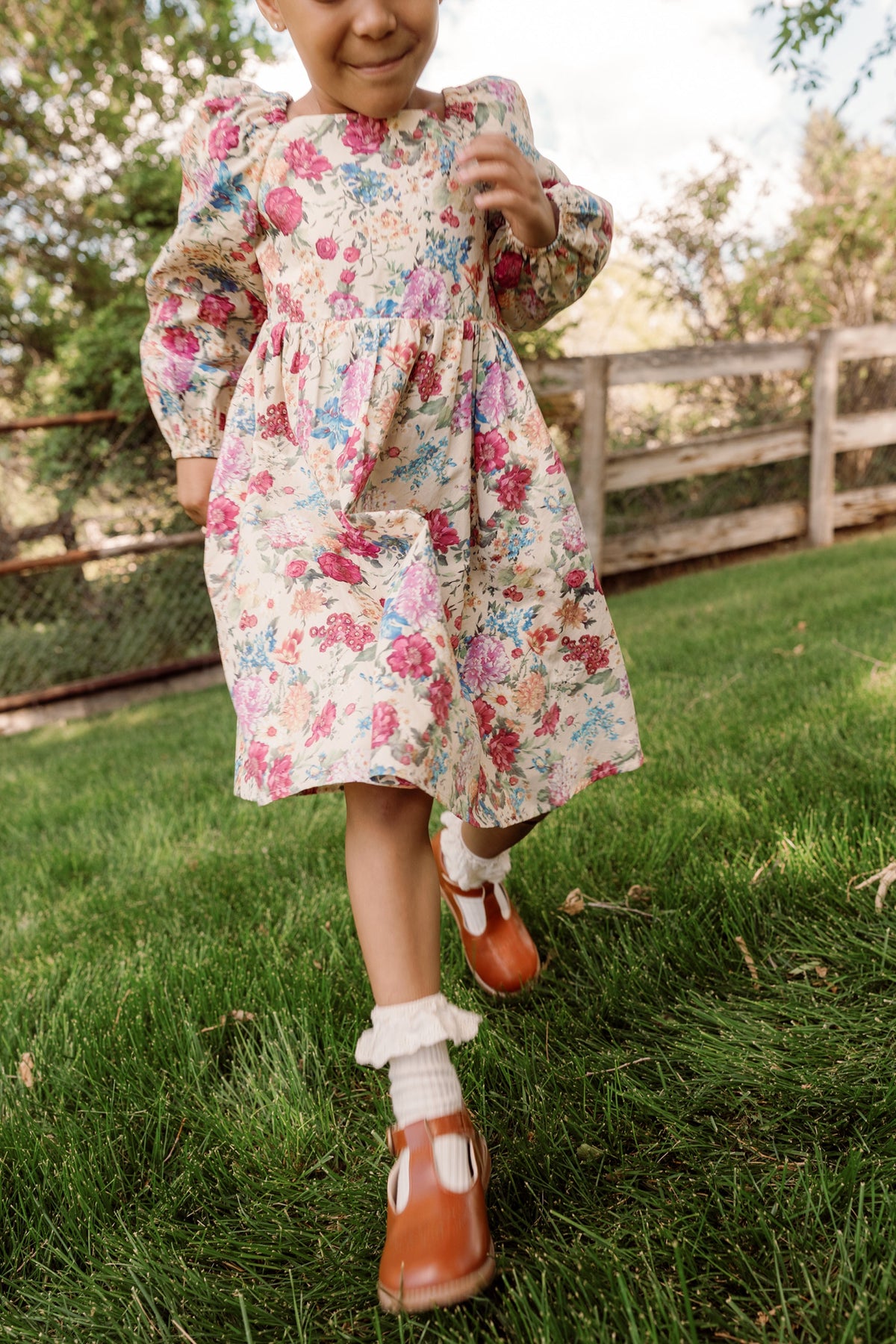 A child wearing the Mini Adelpha Dress in Autumn Amity Floral by Ivy City Co, with its smocked bodice and charming balloon sleeves, smiles while walking on green grass, with a wooden fence and trees in the background.