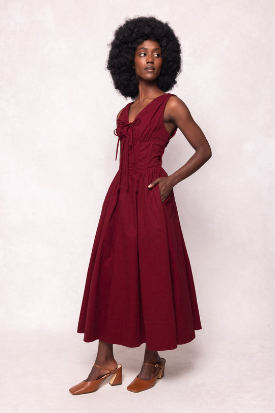 A woman stands in profile against a light background, her hands slipped into the pockets of the Kayla Dress in Burgundy by Ivy City Co—a sleeveless, midi-length dress featuring front ties and a corset waist. She completes her look with brown heeled shoes and wears her natural afro hairstyle.