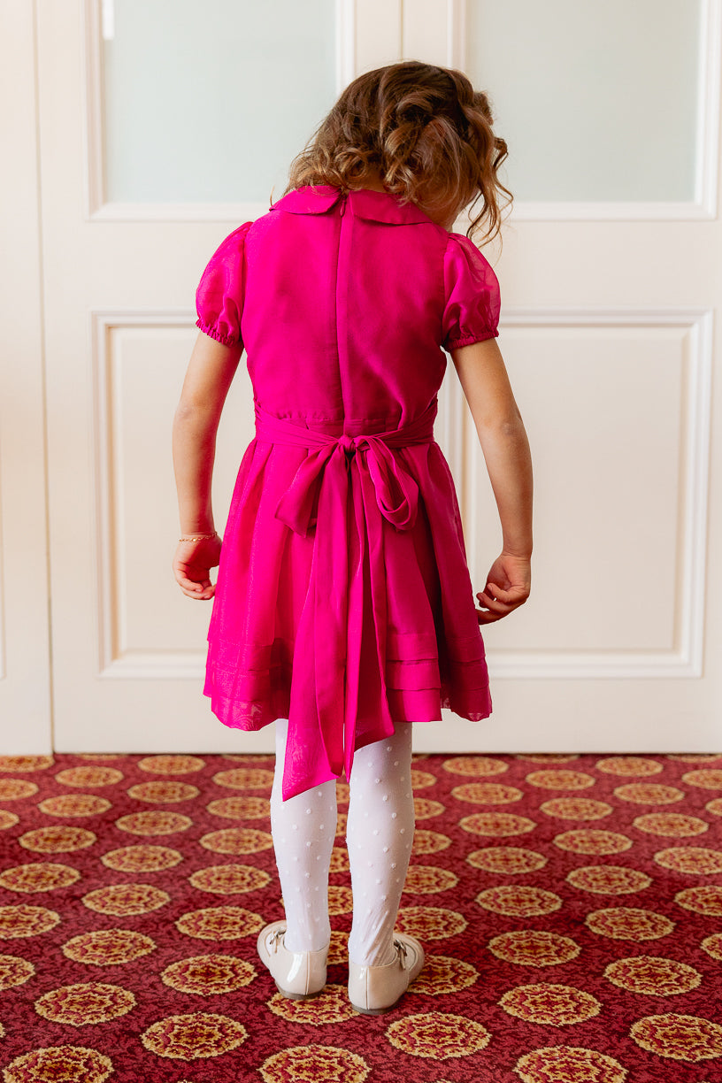 A young girl, dressed in the Ivy City Co Mini Cassian Dress in Hot Pink, stands on a patterned carpet with her back to the camera, facing white double doors. Her look is completed with dotted white tights and cream shoes, making her the picture of charm in this delightful girls’ party dress.