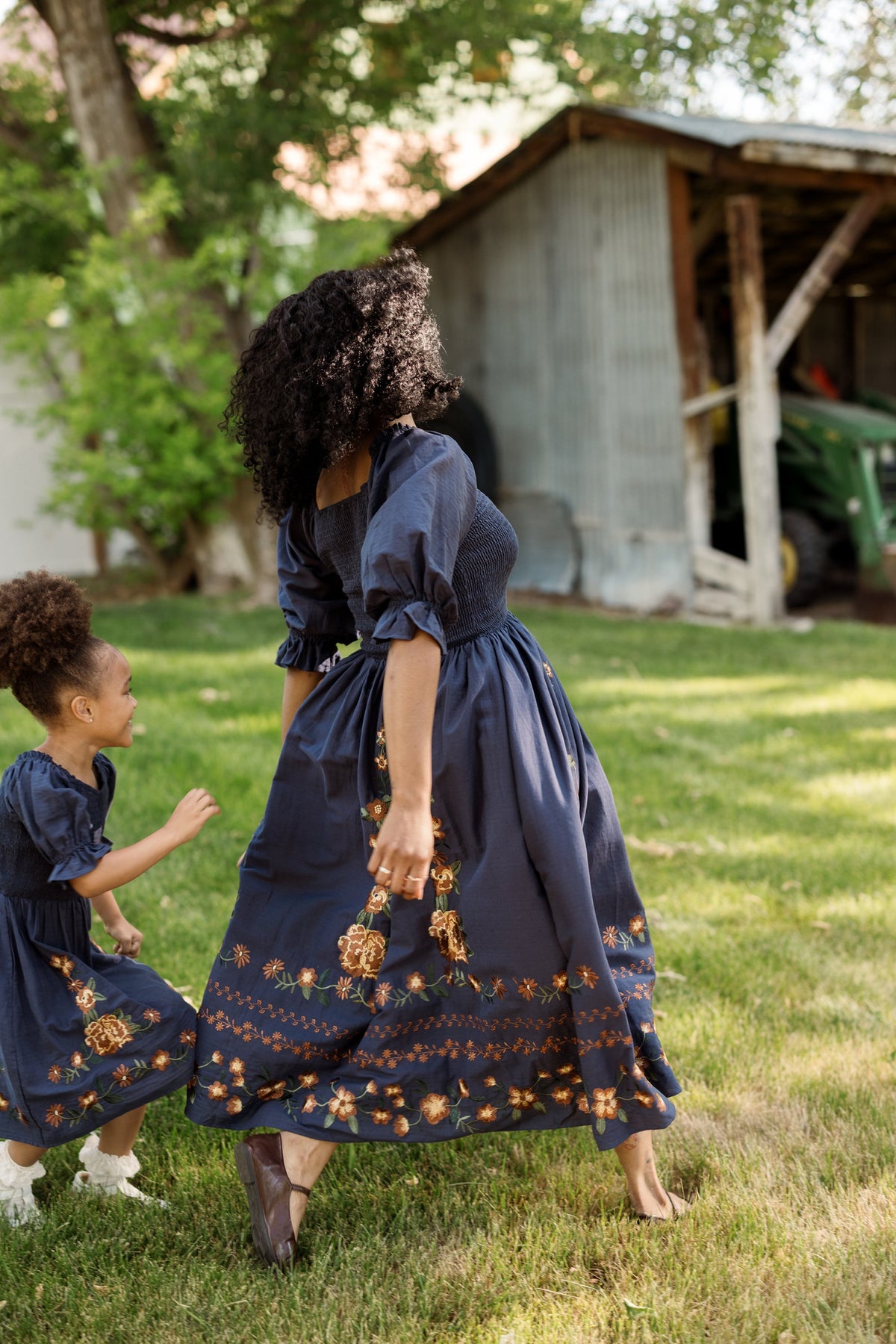 A woman and a young girl, both in matching Ivy City Co Mini Teagan Dresses in Navy Embroidered Fall Floral, stroll and play together on green grass near a rustic shed.