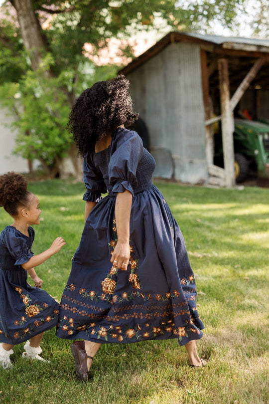 A woman and a young girl, both in matching Ivy City Co Mini Teagan Dresses in Navy Embroidered Fall Floral, stroll and play together on green grass near a rustic shed.