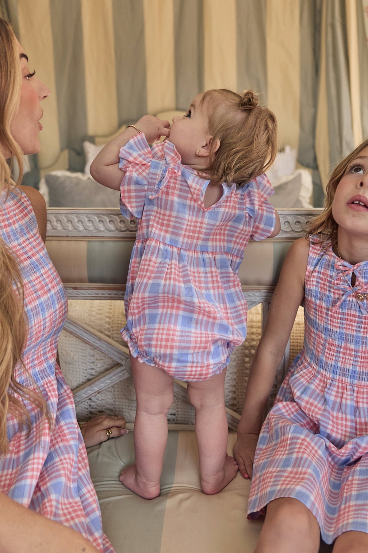 A baby wearing the Ivy City Co Baby Ana Romper in Blossom Plaid stands on a couch between two women dressed in matching plaid dresses. All three are styled in coordinated pastel-colored outfits and are seated indoors.