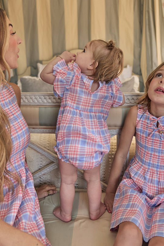 A baby wearing the Ivy City Co Baby Ana Romper in Blossom Plaid stands on a couch between two women dressed in matching plaid dresses. All three are styled in coordinated pastel-colored outfits and are seated indoors.