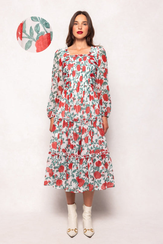 A woman models the Ivy City Co Selena Dress in Red Roses—a long, tiered chiffon midi dress featuring a red and green floral print, long sleeves, and white ankle boots. An inset provides a close-up of the red rose pattern on the dress. The background is plain and light-colored.