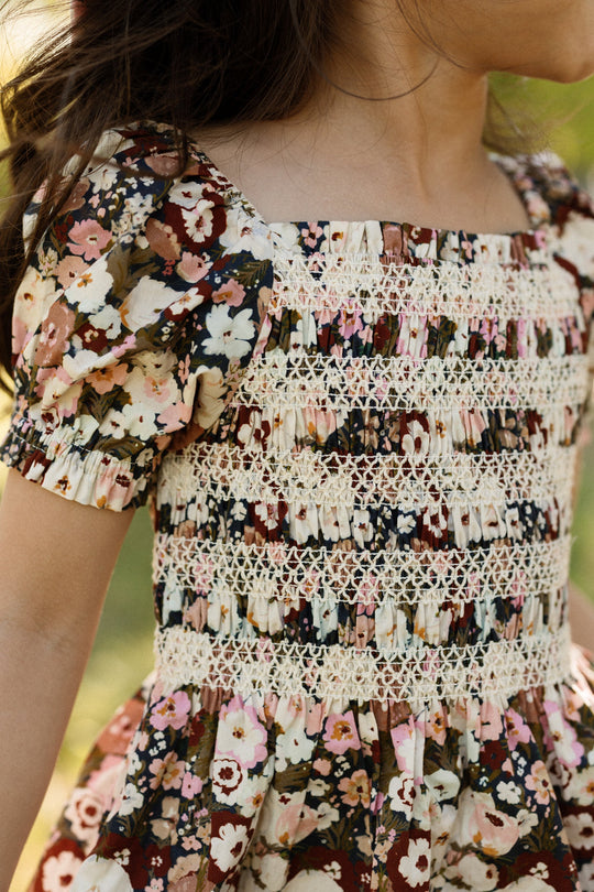 A child in the Ivy City Co Mini Sunday Best Dress in Midnight Garden, featuring puff sleeves, a smocked bodice, and decorative lace details, stands outdoors facing sideways.