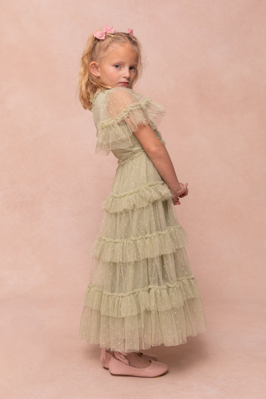 A young girl with blonde hair styled in pink bows wears the Ivy City Co Mini Whimsical Maxi Dress in Sage—featuring a light green ruffled tulle bodice and a layered, polka-dot tulle skirt—paired with pink shoes. She stands sideways against a pale pink background and looks over her shoulder.