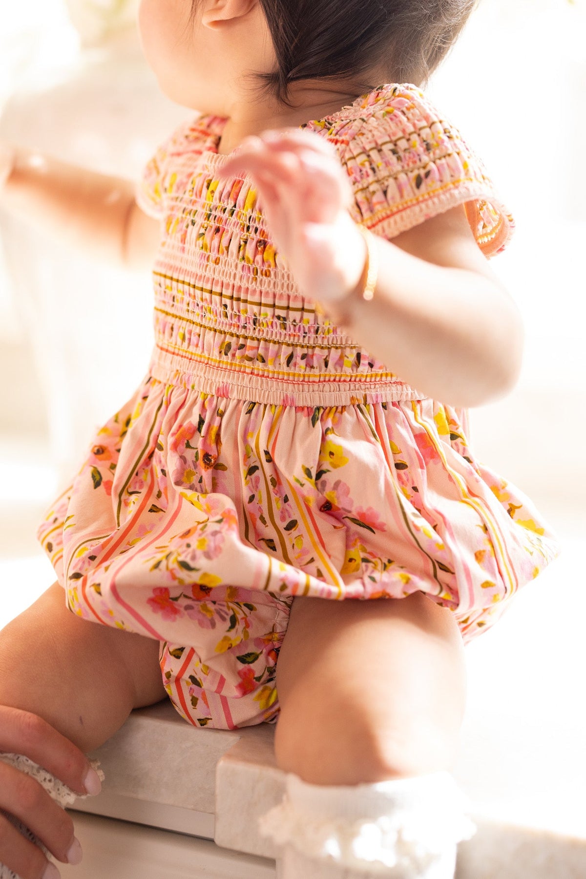 A baby wearing the Ivy City Co Baby Marlow Romper in Peach Handpainted Flora Stripe and white socks sits on a ledge, looking to the side with one arm outstretched as sunlight softly illuminates the scene.