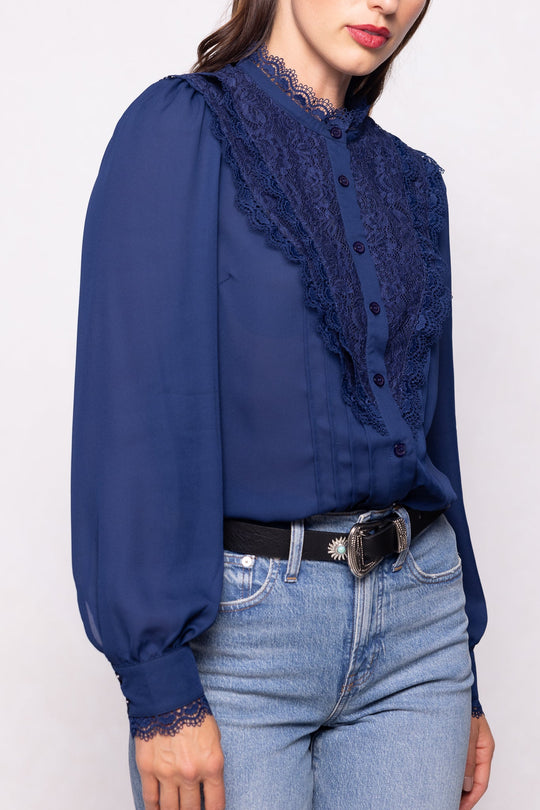 A woman wearing the Ivy City Co Blythe Blouse in Navy—with long lace sleeves—paired with blue jeans, partially tucked in, and accessorized with a black belt featuring a silver buckle. This nursing-friendly look is both stylish and practical. Her face is partially visible.