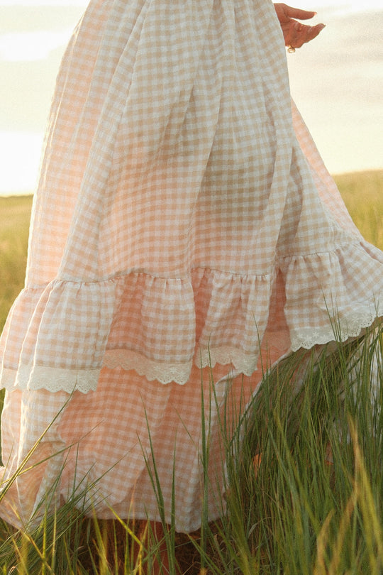A person stands in a sunlit grassy field wearing the Ivy City Co Presley Sleeveless Dress in Cream Gingham; only the lower half of the maxi-length, ruffled dress and one hand are visible.