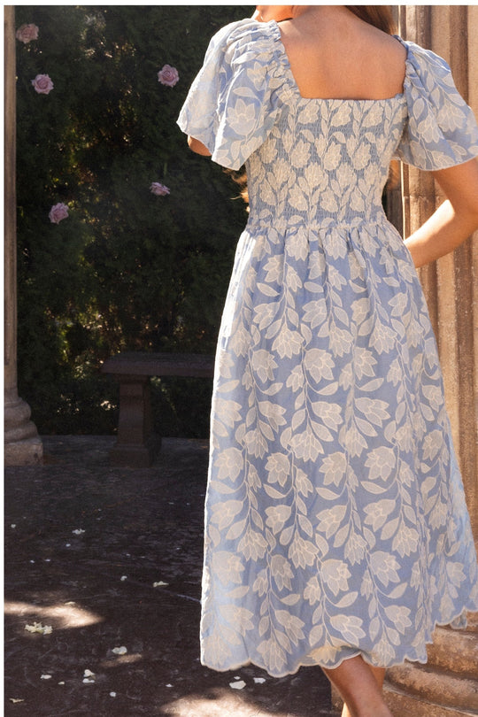 Wearing the Ophelia Dress in Mosaic Embroidery by Ivy City Co, a person stands outdoors with their back to the camera, surrounded by stone columns and lush greenery.