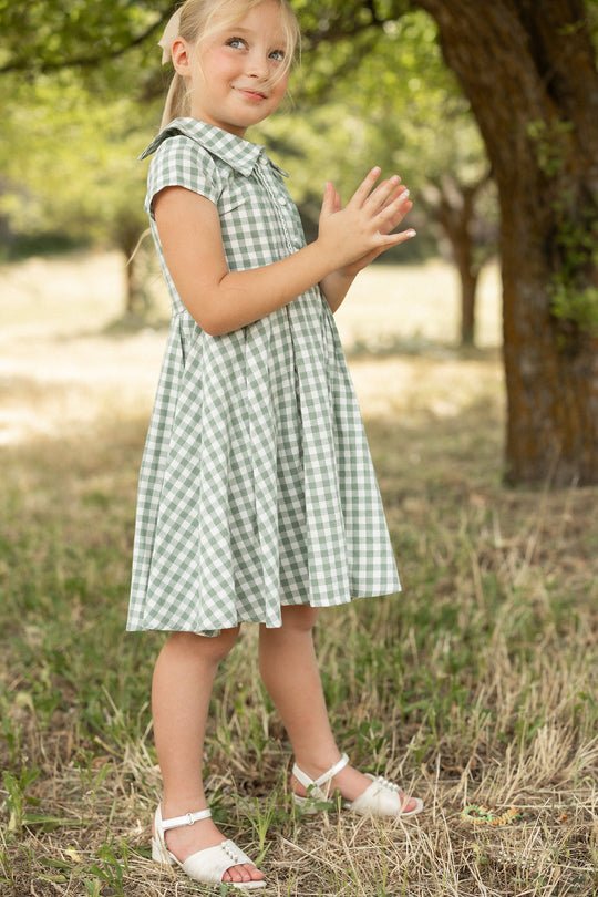 A young girl stands outside on the grass near a tree on a sunny day, smiling with her hands together while wearing the Ivy City Co Mini Amber Dress in Sage Gingham.