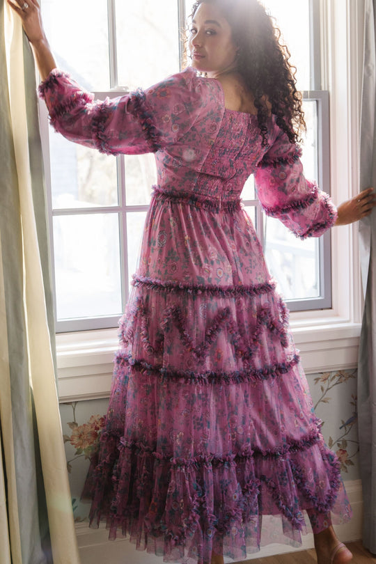 Wearing the Ivy City Co Catherine Dress in Climbing Vine Bloom, a person with curly hair stands by a window, looking over their shoulder as soft light streams in.