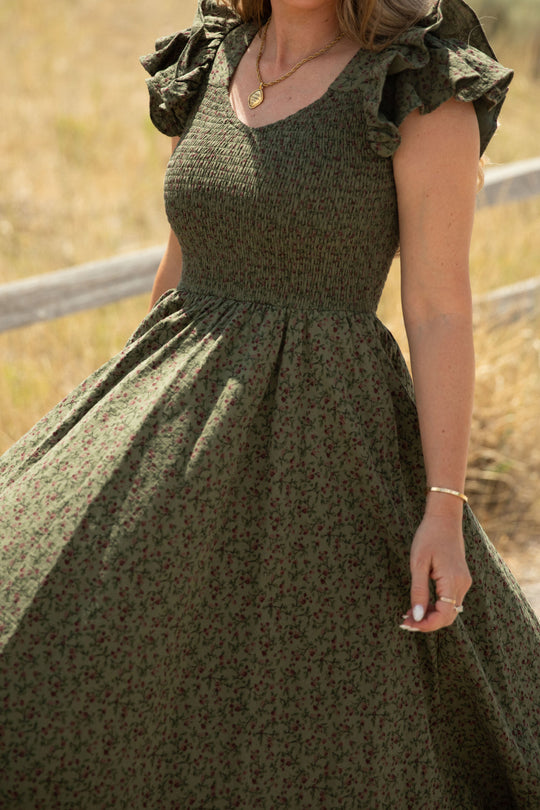 A woman wearing the Hattie Dress in Woodland Floral by Ivy City Co—a green, floral-patterned style featuring ruffled sleeves and a smocked bodice—stands outdoors near a wooden fence. She accessorizes with a gold necklace and bracelet, and her dress billows gracefully as she holds the skirt, offering a charming and maternity-friendly look.