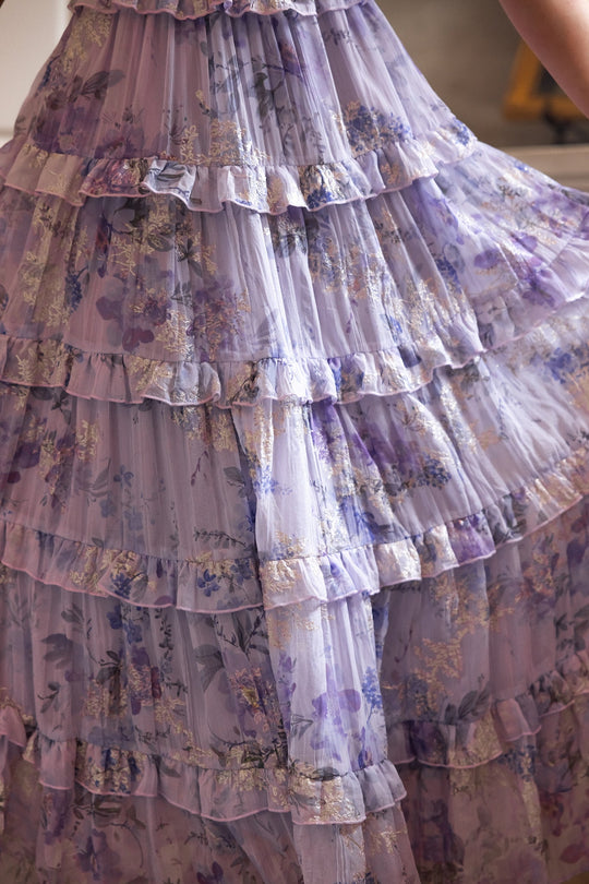 A close-up of the Ivy City Co Winnie Dress in Lavender Aquarelle Fleur showcases its tiered, ruffled chiffon design with a floral pattern and sheer fabric. Soft lavender hues and subtle gold accents make this maternity-friendly dress both elegant and comfortable.
