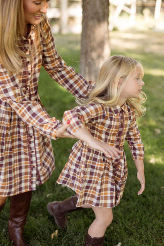 A woman and a young girl are outdoors on grass, both wearing matching Ivy City Co Mini Bowie Long Sleeve Dresses in Spiced Plaid with balloon sleeves and brown boots. The woman reaches for the girl, who looks to the side while in motion.