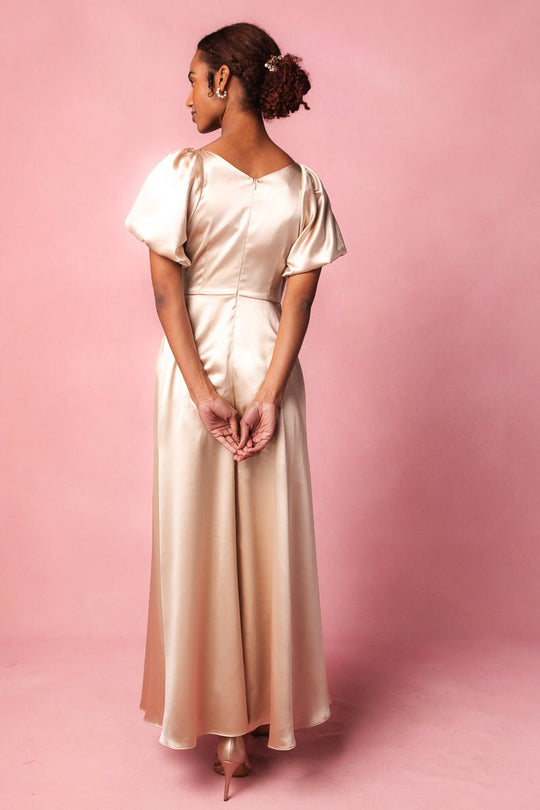 A woman wearing the Tessie Dress in Champagne by Ivy City Co stands on a pink background, facing away with her hands clasped behind her back. Her hair is styled in a curly low bun, showcasing this elegant satin dress—an ideal choice for a bridesmaid or a stylish wedding guest.