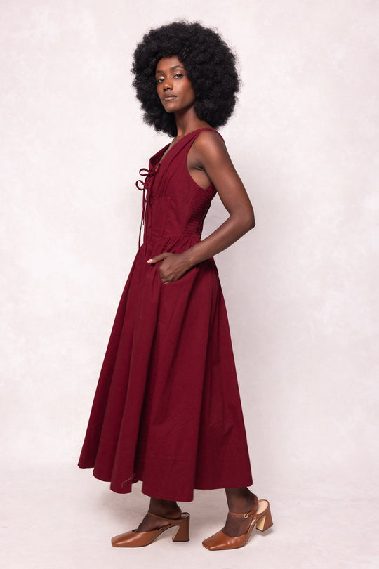 A woman with an afro hairstyle stands sideways against a light background, wearing the Ivy City Co Kayla Dress in Burgundy—a sleeveless midi dress featuring lace-up detailing and a flattering corset waist—paired with brown heeled shoes.