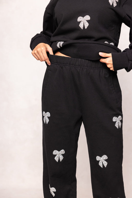 A person is shown from shoulders to ankles, adjusting the waistband of the Ivy City Co Micah Jogger Sweatpants in Black, which feature glittery rhinestone bow designs. The background is plain and light-colored.