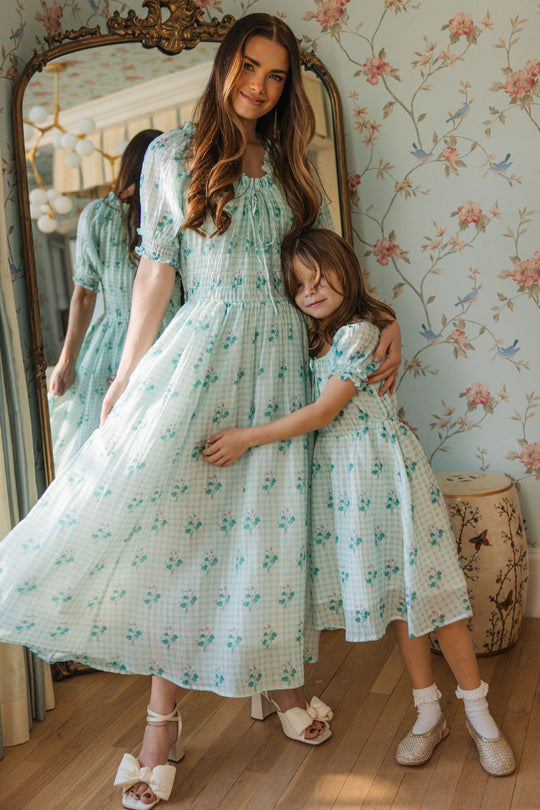 A woman and a young girl smile together in front of a large ornate mirror in a softly lit room with floral wallpaper, both wearing the Ivy City Co Jessie Dress in Bloom Gingham. Their charming matching outfits are maternity friendly, perfect for mothers and daughters to share special moments. The girl lovingly hugs the woman’s waist.