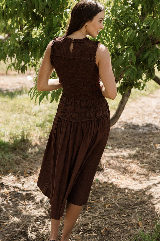 A woman stands outdoors on dry grass near a leafy tree, facing away in warm sunlight and wearing the Ivy City Co Simone Dress in Warm Cocoa—a brown, sleeveless dress with a smocked bodice that evokes a natural, summery atmosphere.