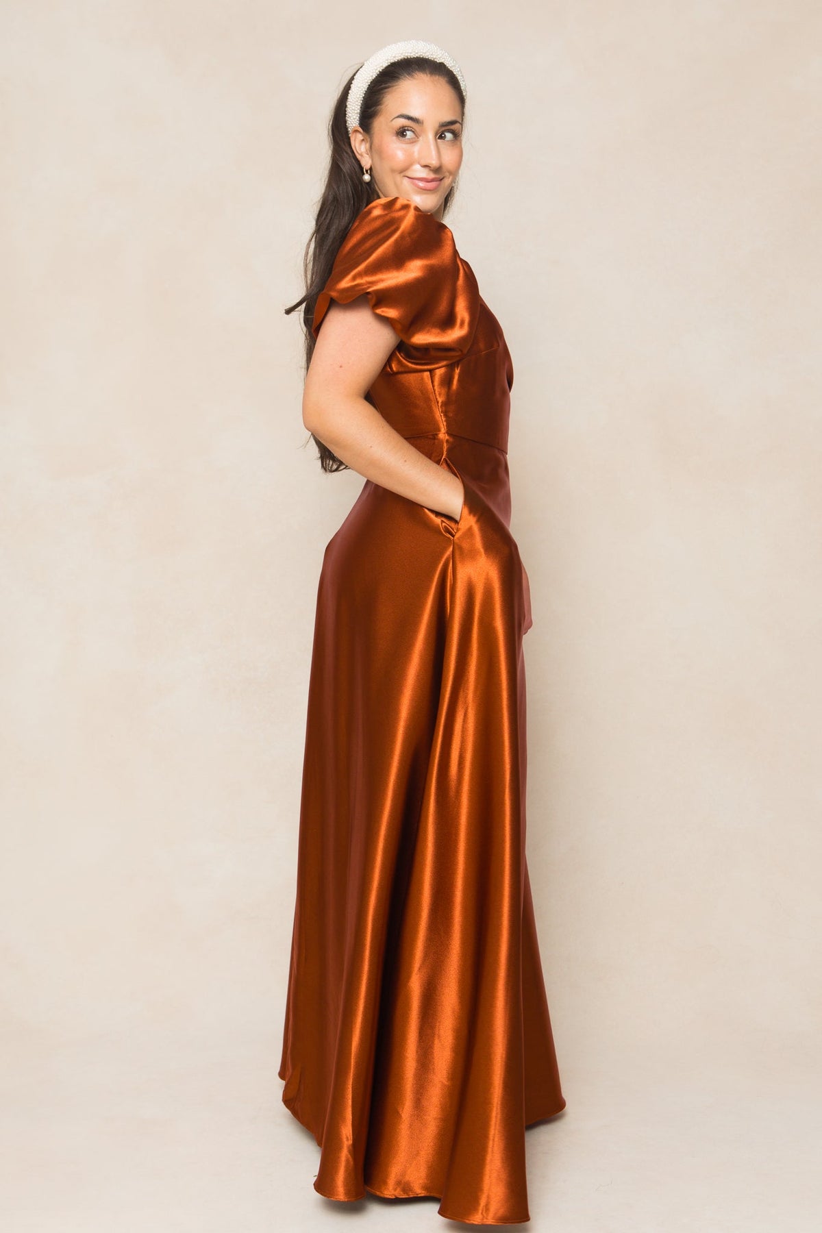 A woman stands sideways and smiles against a light beige background, wearing the Ivy City Co Tessie Dress in Amber Satin—a nursing-friendly dress with short sleeves and pockets.