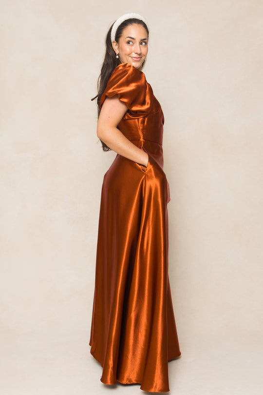 A woman stands sideways and smiles against a light beige background, wearing the Ivy City Co Tessie Dress in Amber Satin—a nursing-friendly dress with short sleeves and pockets.