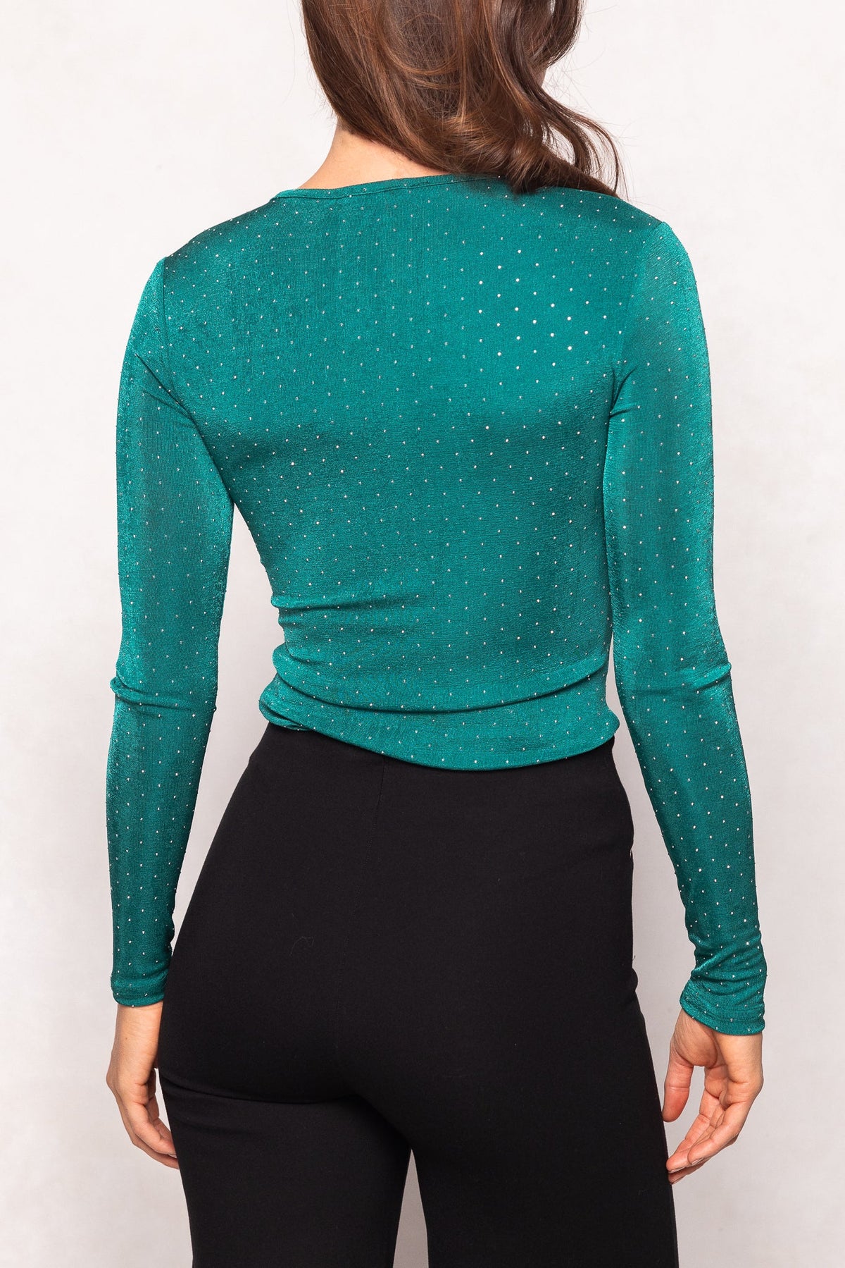 A person with long brown hair is shown from the back wearing the Ivy City Co Bravo Top in Emerald—a fitted, long-sleeve top adorned with jewel embellishments and small shiny details—paired with black high-waisted pants.