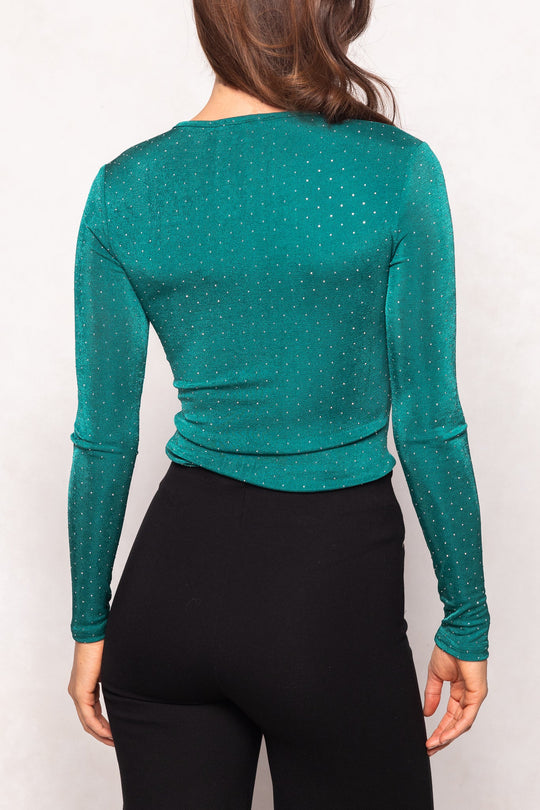 A person with long brown hair is shown from the back wearing the Ivy City Co Bravo Top in Emerald—a fitted, long-sleeve top adorned with jewel embellishments and small shiny details—paired with black high-waisted pants.