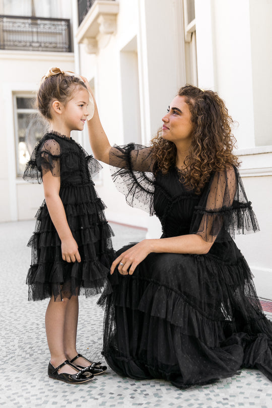 A woman kneels outside a white building, gently touching the head of a young girl dressed in Ivy City Co’s Mini Whimsical Dress in Black, which features a delicate layered tulle skirt. The woman is also wearing a long black dress, matching the girl's look.