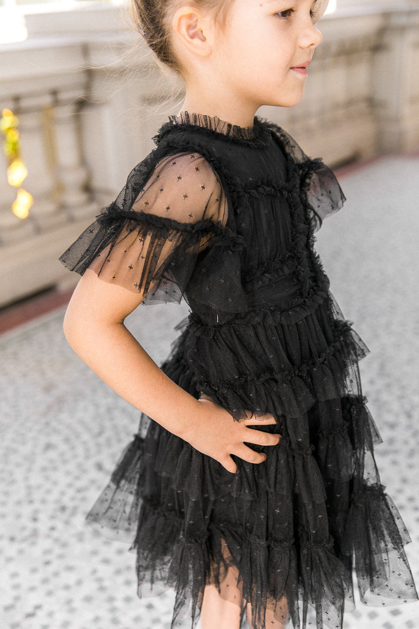 A young girl stands in profile with one hand on her hip, outdoors near a decorative stone railing, wearing the Ivy City Co Mini Whimsical Dress in Black. The short-sleeved dress features ruffled, sheer layers and star patterns, while its layered tulle skirt adds playful charm to her look.