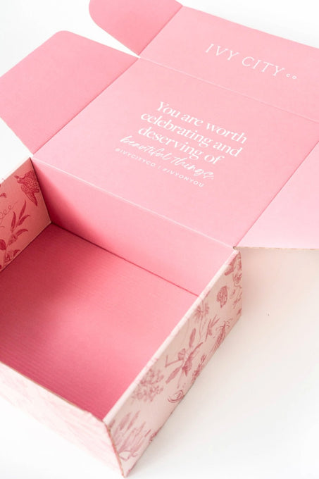 An open pink Ivy City Co Gift Box with floral sides and a message inside the lid: You are worth celebrating and deserving of beautiful things. Brand name is printed above the message.