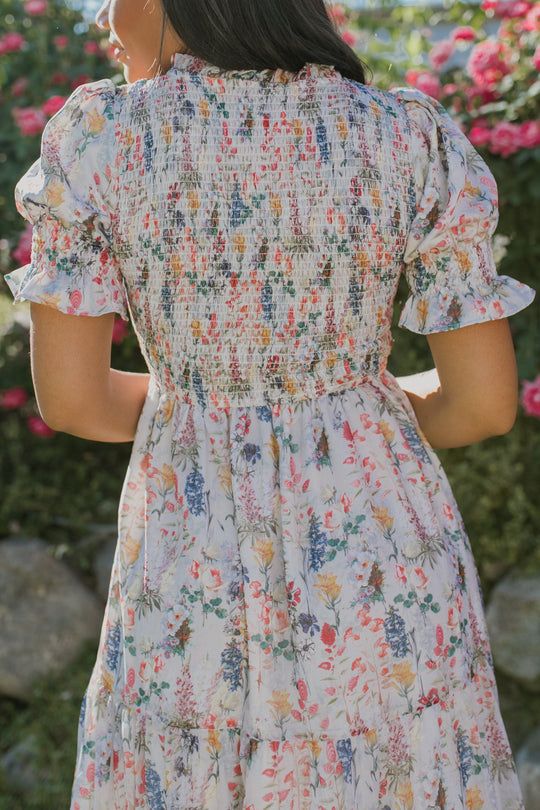 A woman stands outdoors with her back to the camera, wearing the Ivy City Co Cordelia Floral Dress, a maternity-friendly style with a smocked bodice. Pink flowers and greenery surround her in the background.