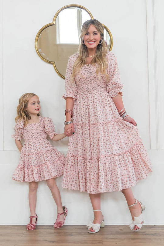 A woman and a young girl smile, holding hands in matching Ivy City Co Madeline Dress In Pink—maternity-friendly with floral smocking—paired with white sandals, in a bright room with white walls and a gold-framed mirror.