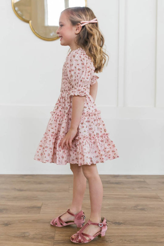 A young girl with wavy blonde hair smiles sideways in the Ivy City Co Mini Madeline Dress in Pink, featuring ruffles and a smocked bodice, paired with pink sandals and a matching hair clip, standing on a wooden floor by a white wall with a round mirror.