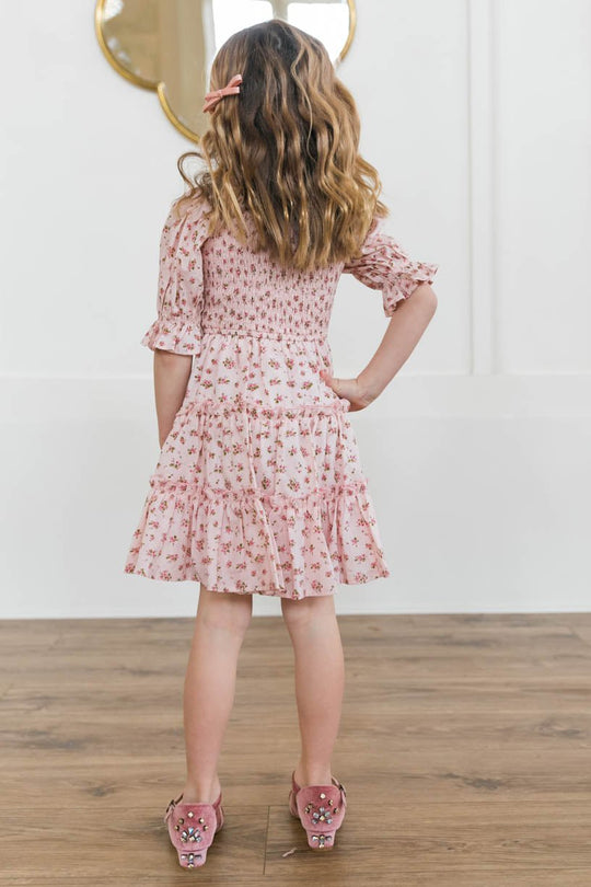 A young girl with wavy, light brown hair wears the Mini Madeline Dress in Pink by Ivy City Co, featuring a smocked bodice, as she stands on a wooden floor facing away from the camera.
