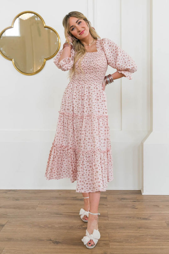 A woman in the Ivy City Co Madeline Dress in Pink, a floral tea-length, maternity-friendly dress with puff sleeves, stands indoors on a wooden floor. She smiles with one hand on her head, wearing white sandals and silver bangles by a gold-framed mirror.