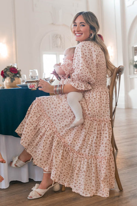 A woman in the maternity-friendly Madeline Dress In Pink by Ivy City Co sits indoors at a table, smiling and holding a baby on her lap amid elegant decor and natural light.