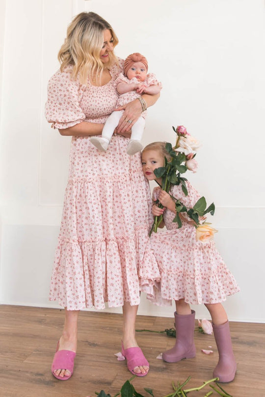 A woman, baby, and young girl wear matching Ivy City Co Mini Madeline Dresses in Pink. The girl stands in purple boots, holding flowers, with more blossoms scattered across the wooden floor.
