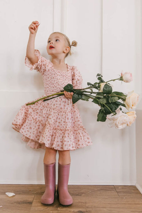 A young girl wears the Ivy City Co Mini Madeline Dress in Pink, holding pale roses and reaching upward. She stands against a white wall and wooden floor, her purple rain boots adding a playful touch.