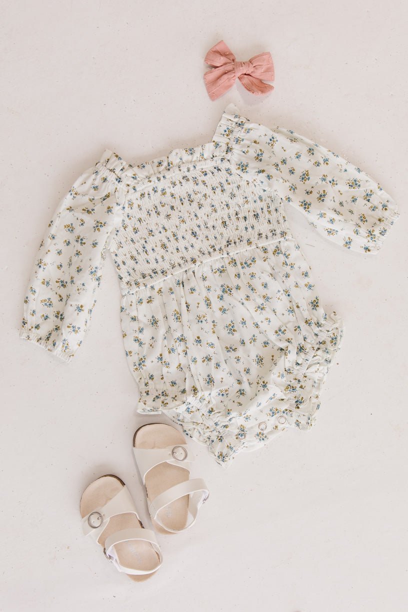 Flat lay featuring the Baby Madeline Romper in Blue by Ivy City Co, paired with cute white buckle sandals and a pink bow, all beautifully arranged on a light background.