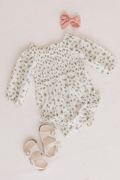 Flat lay featuring the Baby Madeline Romper in Blue by Ivy City Co, paired with cute white buckle sandals and a pink bow, all beautifully arranged on a light background.