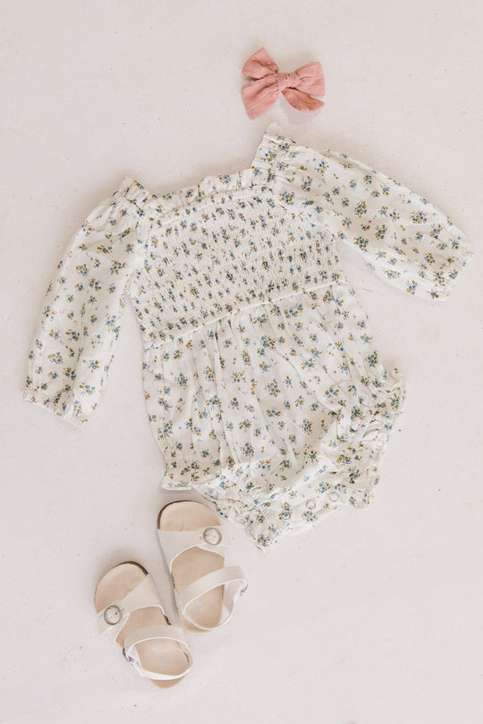 Flat lay featuring the Baby Madeline Romper in Blue by Ivy City Co, paired with cute white buckle sandals and a pink bow, all beautifully arranged on a light background.