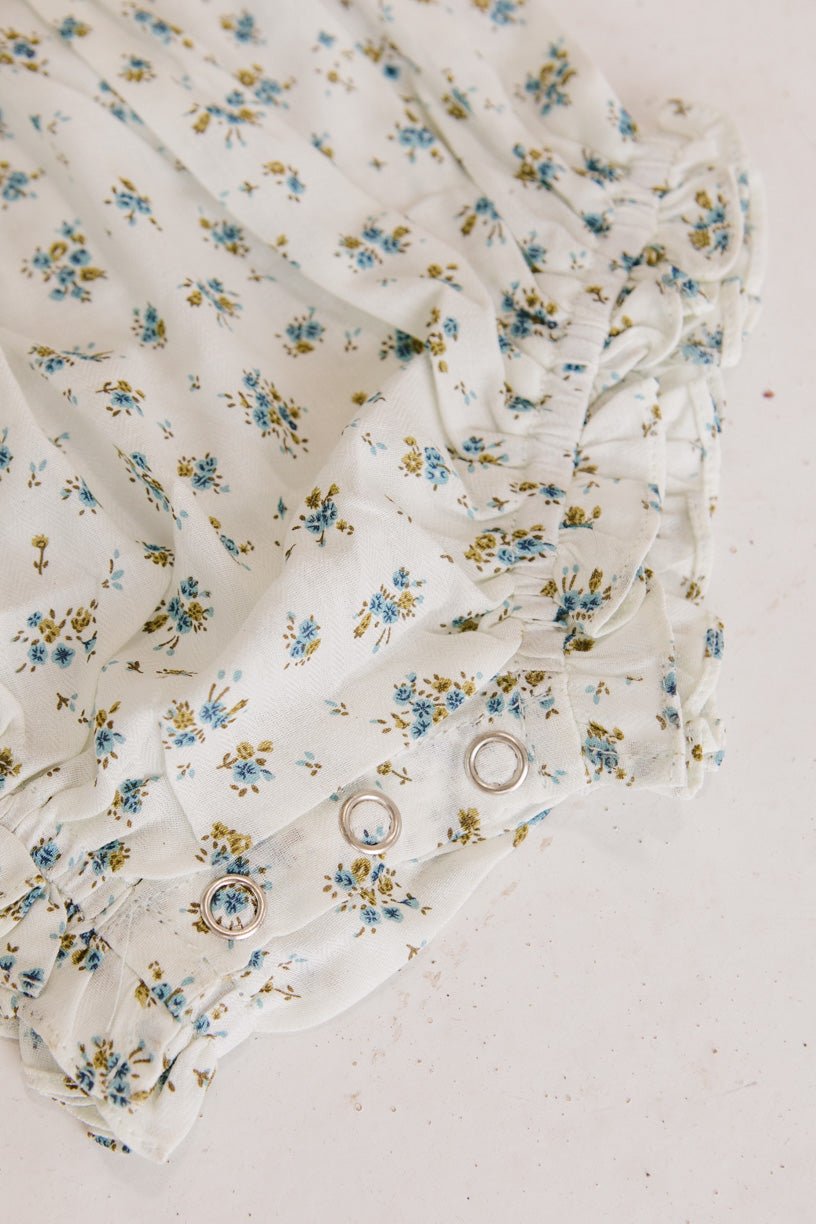 Close-up of Ivy City Cos Baby Madeline Romper in Blue, a white floral romper with blue and yellow flowers, ruffles, puff sleeves, and silver snap buttons—ideal for babies as a cute outfit or for special occasions.