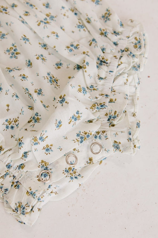 Close-up of Ivy City Cos Baby Madeline Romper in Blue, a white floral romper with blue and yellow flowers, ruffles, puff sleeves, and silver snap buttons—ideal for babies as a cute outfit or for special occasions.